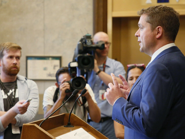 Texas Gouverneur Greg Abbott im Anzug und Krawatte steht an einem Podium mit Papieren und Mikrofonen, während er eine Gruppe von Menschen anspricht, von denen einige Kameras halten, mit einer gerahmten Wand im Hintergrund.