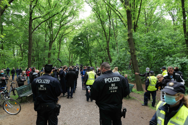 Polizeibeamte vor einer Menge bei einer Anti-Terror-Demonstration in Berlin, mit Fahrrädern und einer Bank im Vordergrund und Bäumen im Hintergrund.