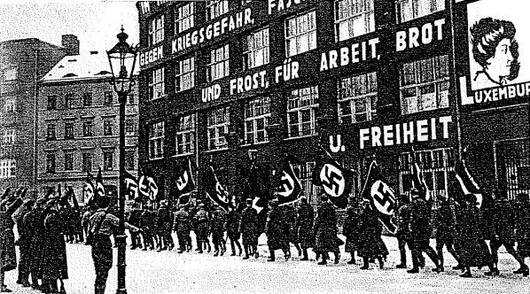 Schwarzes und wei├čes Bild einer Gruppe, die mit Fahnen und Transparenten die Stra├če entlangmarschiert, mit einem Lichtmast links und einem Geb├Ąude mit der Aufschrift "Deutsche Nationalsozialistische Arbeiterpartei in Berlin" rechts.