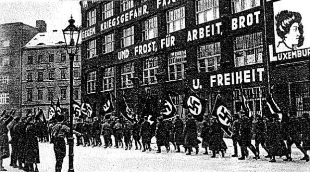 Schwarzes und wei├čes Bild einer Gruppe, die mit Fahnen und Transparenten die Stra├če entlangmarschiert, mit einem Lichtmast links und einem Geb├Ąude mit der Aufschrift "Deutsche Nationalsozialistische Arbeiterpartei in Berlin" rechts.