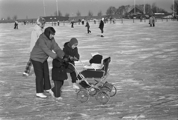 Schwarzes und weißes Bild von Menschen, die auf einer Eisbahn Schlittschuh laufen, mit einem Baby im Kinderwagen im Vordergrund, Pfosten, Bäume, Häuser und einen klaren Himmel im Hintergrund.