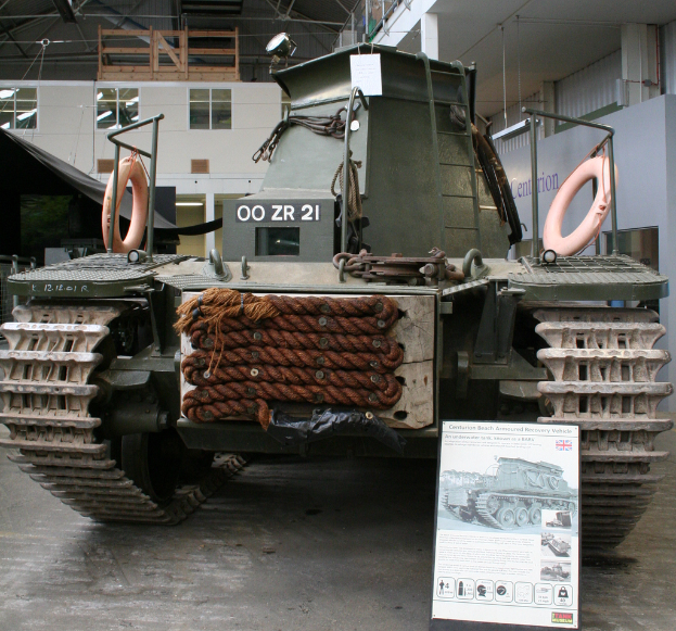 Ein Panzerfahrzeug ist auf dem Boden positioniert mit einer Tafel und mehreren Bildern in der Nähe, vor einer Wand, Säulen, Lampen, einem Schuppen und Stangen.