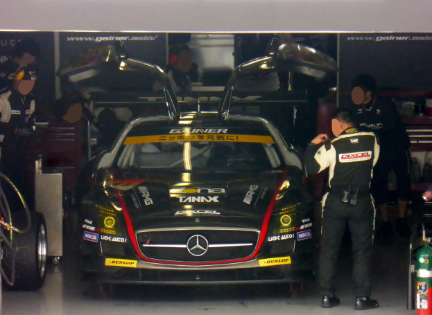 Ein silberner Mercedes-Benz SLS AMG GT3 Rennauto in einer Garage mit zwei Personen davor, einem Feuerlöscher rechts und Informationsschildern im Hintergrund.