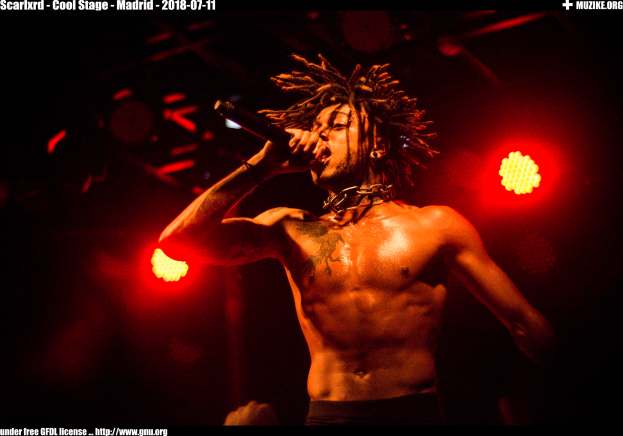 Ein Mann mit Dreadlocks singt ohne Hemd auf der Bühne in ein Mikrofon, mit Text oben und unten, der "scarikrd - cooler Stage - madrid 2018-07-11" lautet.