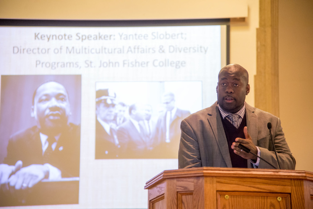 Ein Mann in einem Mantel, Krawatte und Hemd spricht rechts neben einem Podium, mit einem Projektionsschirm in der Mitte und einem Mann in Schwarz und Weiß links.