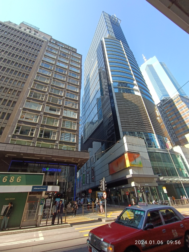 Ein rotes Auto fährt eine von hohen Wolkenkratzern gesäumte Stadtstraße entlang, mit Passanten, Verkehrsampeln und Beschilderung, unter einem klaren Himmel.