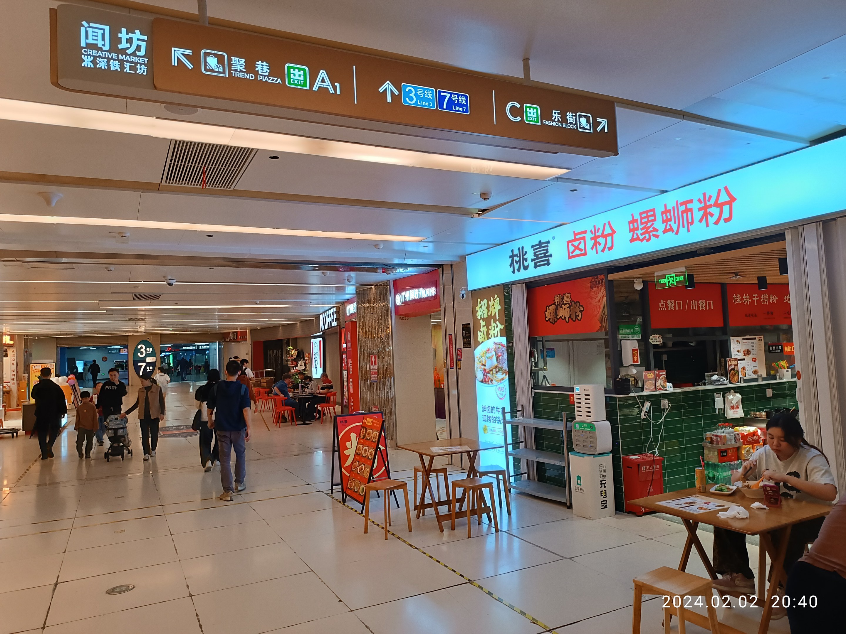 Eine Gruppe von Menschen, die durch ein Einkaufszentrum geht, mit Tischen, auf denen verschiedene Gegenstände ausgelegt sind, Schilder an den Wänden, Deckenleuchten und einem zentralen kreativen Markt mit Essensauslagen.