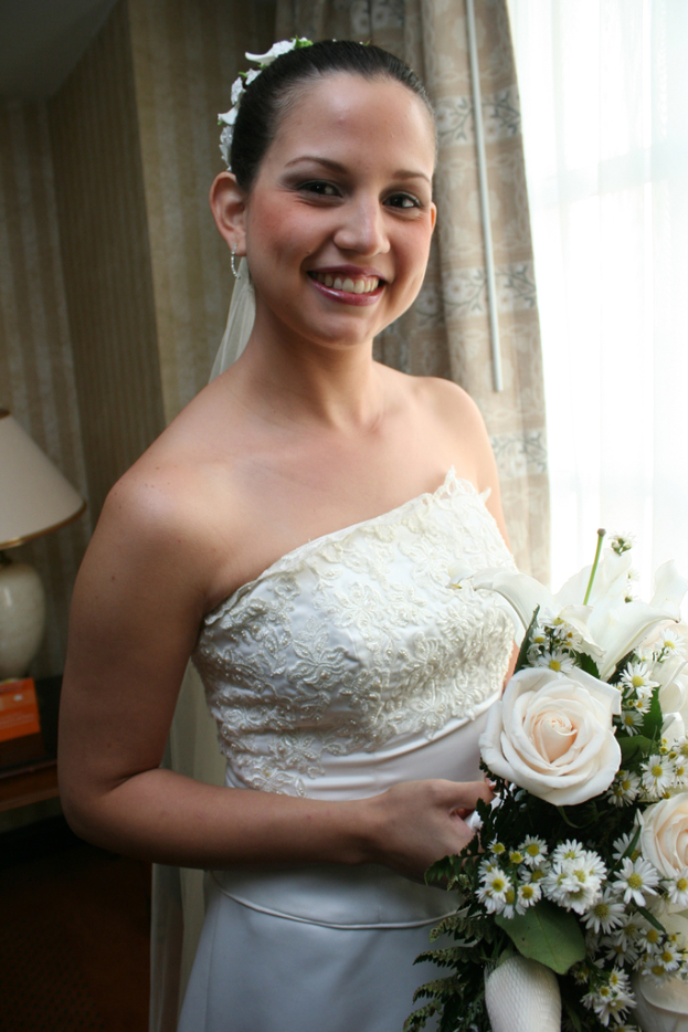 Eine Braut steht lächelnd, hält einen Strauß, mit einem Vorhang und einer Wand im Hintergrund und einer Lampe auf einem Tisch links.