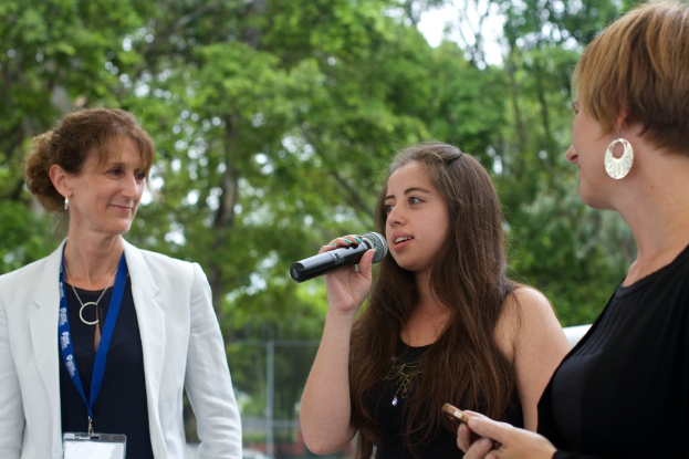Drei Frauen, eine singt in ein Mikrofon, eine andere lacht und trägt eine Anstecknadel, mit einem Geländer, einem Baum und Himmel im Hintergrund.