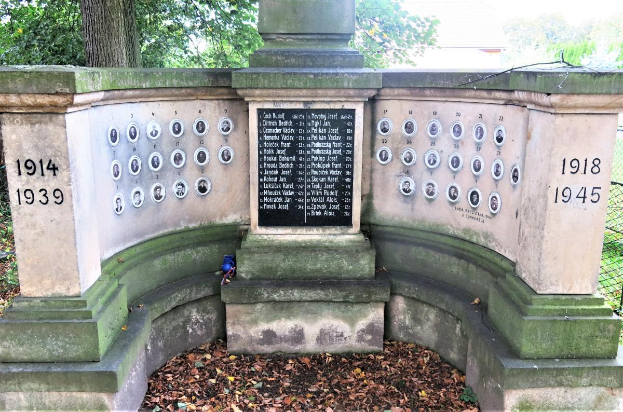 Ein Denkmal in einem von Bäumen umgebenen Park mit einem Zaun, einer Tafel mit Text und Zahlen sowie trockenen Blättern auf dem Boden, das sich im Jüdischen Friedhof in Berlin befindet und als Gedenkstätte für Holocaust-Opfer dient.