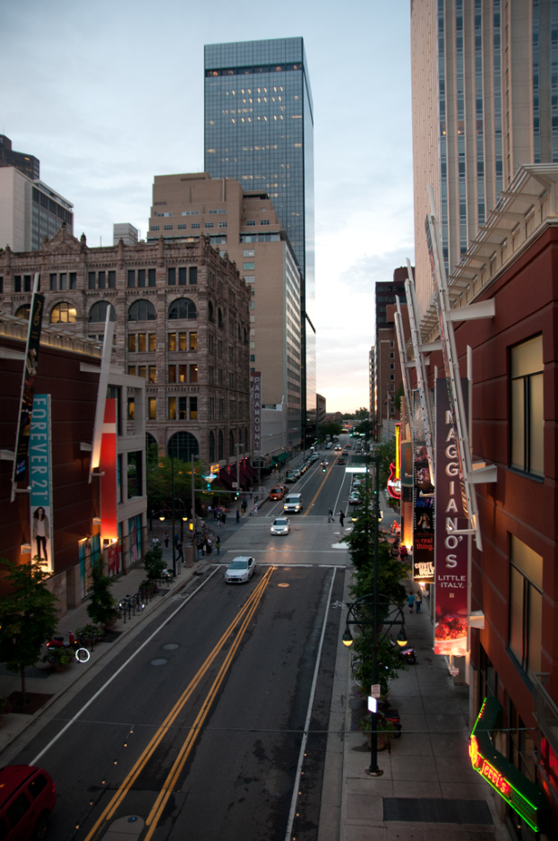 Eine Stadtansicht mit einer von Autos befahrenen Straße unten, flankiert von Gebäuden und Wolkenkratzern auf beiden Seiten, unter einem Himmel oben.