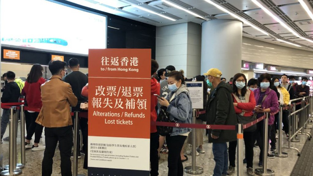 Menschen mit Masken stehen in einer Schlange am Hong Kong Airport, getrennt durch Barrierepfosten mit Bändern, mit einem Rückerstattungs-Schild und Deckenleuchten im Hintergrund.