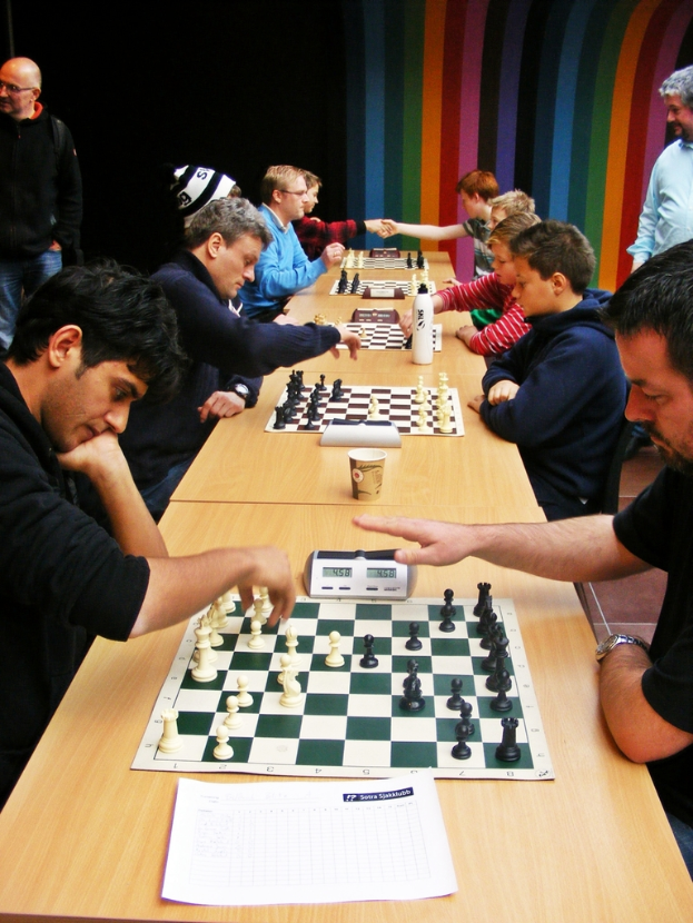 Zwei Personen sitzen sich gegenüber und spielen Schach auf einem Brett, mit einem Timer auf dem Tisch.