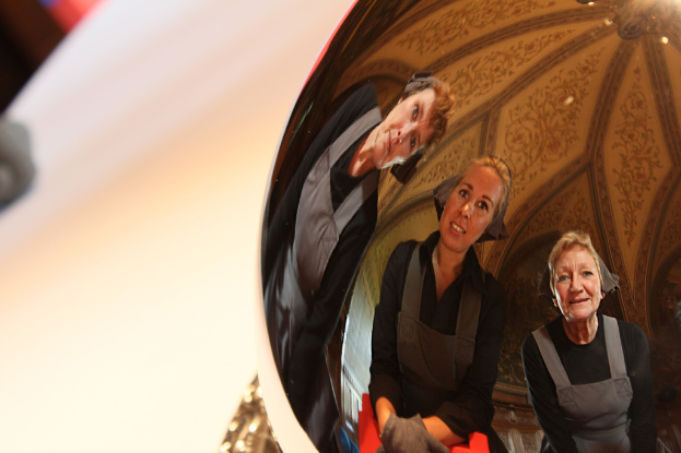 Ein reflektierender Ball auf einem weißen Tisch zeigt das Spiegelbild von drei lächelnden Frauen, die sich leicht biegen, mit einem Dach im Hintergrund.