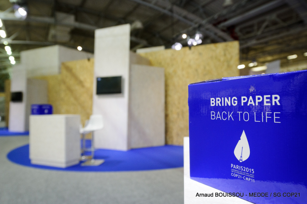 Ein Messestand mit einem blauen und weißen "Bring Paper Back to Life"-Schild auf der rechten Seite, mit unscharfen Stühlen, Tischen, einem wandmontierten Fernseher und Deckenlampen im Hintergrund.