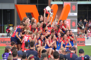 Gruppe von Frauen-AFL-Spielerinnen feiert mit einem Pokal vor einem Publikum, trägt Sportuniformen.
