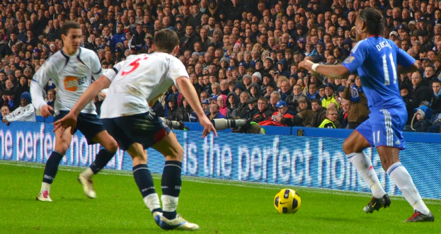 Personen mit einem Ball im Vordergrund, eine Tafel mit Text in der Mitte und Kameras hinter der Tafel sowie eine Gruppe von Personen im Hintergrund.