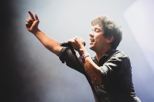 Ein Mann in einem schwarzen Hemd und Jeans singt in ein Mikrofon auf einer Bühne, mit hellen Scheinwerfern und einer jubelnden Menge im Hintergrund.