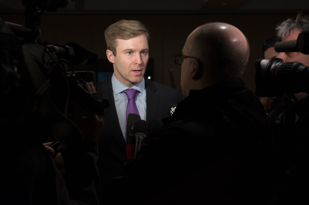 Ein Mann im Anzug und Krawatte, der eine Brille trägt, wird von Reportern interviewt, während zwei Personen mit Kameras rechts und links von ihm stehen und ein Mikrofon vor ihm liegt.