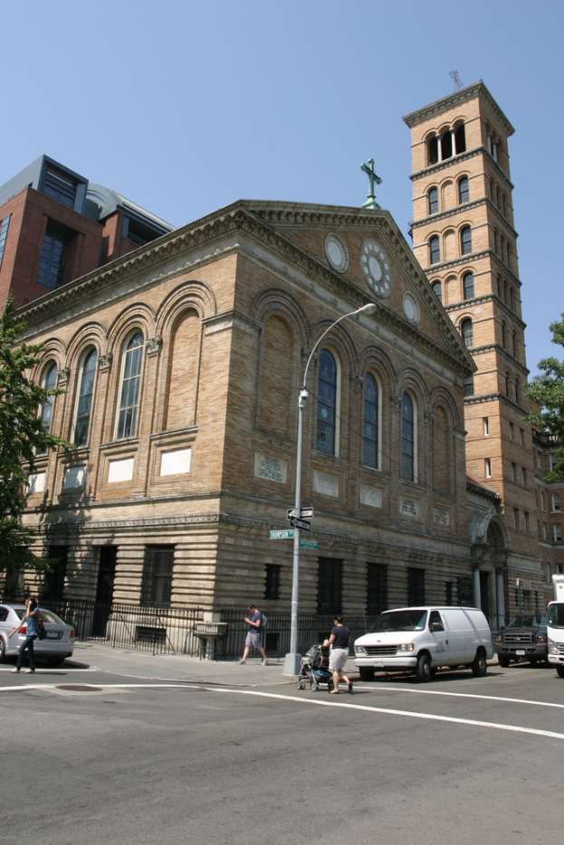 Mehrere Gebäude mit Masten, Bäumen und geparkten Fahrzeugen entlang einer Straße.