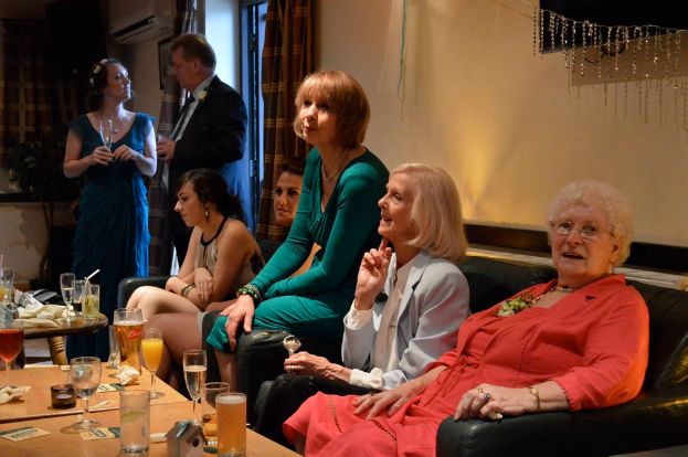 Eine Gruppe von Menschen sitzt und steht in einem Raum mit Tischen, die Gläser, Tassen, Karten und andere Gegenstände halten, einer Tür, Vorhängen, einer Pflanze, einer Klimaanlage und einem Foto auf der Wand sowie einem Fernseher auf der rechten Seite.