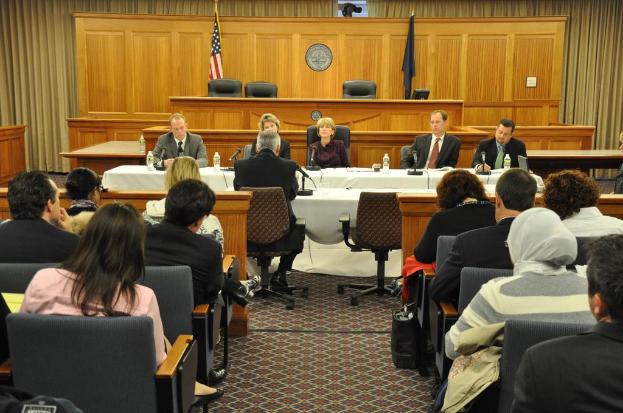 Ein Konferenzsaal mit einer Gruppe von Menschen, die auf Stühlen sitzen, zwei ausländische Flaggen, ein oranger Schreibtisch mit Wasserflaschen und ein Mikrofon.