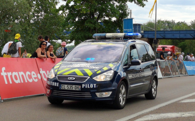 Polizeiauto fährt auf einer Straße neben einer Menschenmenge, mit einem Banner auf der linken Seite des Autos und Geländern mit Bannern dahinter, Bäume und eine Brücke im Hintergrund.