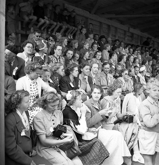 Schwarzes und weißes Foto einer Menge, die in einem Stadion sitzt, mit einer hölzernen Wand und einem Dach im Hintergrund, einige halten Kameras.