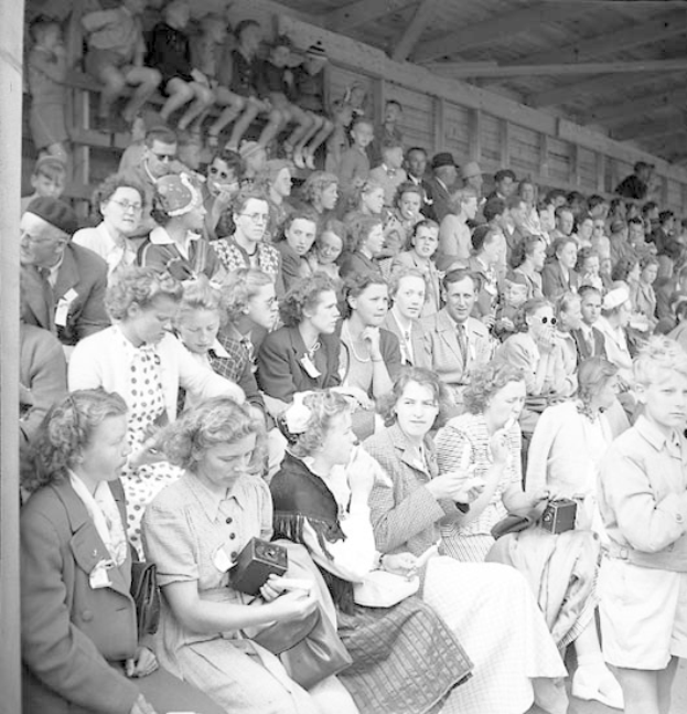 Schwarzes und weißes Foto einer Menge, die in einem Stadion sitzt, mit einer hölzernen Wand und einem Dach im Hintergrund, einige halten Kameras.