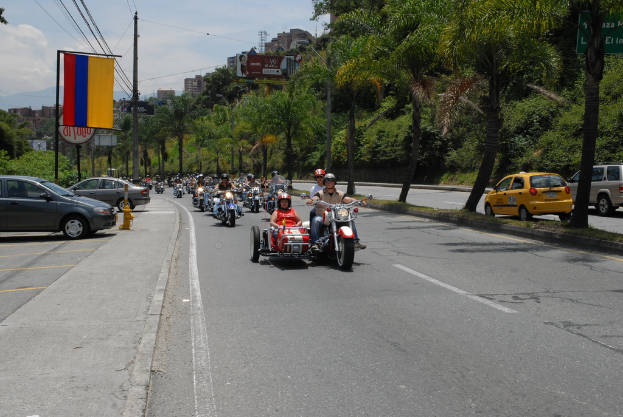 Mehrere Motorräder befinden sich auf einer Straße mit Autos auf beiden Seiten, Gebäuden im Hintergrund und Oberleitungen.