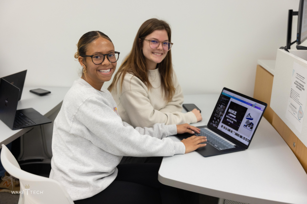 Zwei Frauen mit Brille l├Ącheln beim Arbeiten an Laptops an einem Schreibtisch mit Monitoren, einem Telefon und anderen Gegenst├Ąnden, mit einer Wand im Hintergrund, die darauf hindeutet, dass sie an einem Projekt an der Wake Tech arbeiten.