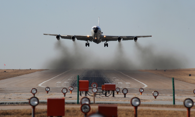 Ein Flugzeug ist oben auf einem Bild positioniert, das eine zentrale Start- und Landebahn zeigt, mit Lampen und Kisten unten und Erde auf beiden Seiten der Start- und Landebahn.