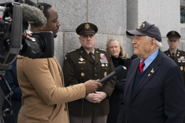 Mann im Anzug und Kappe mit Mikrofon spricht in ein Mikrofon, während ein Reporter ein Objekt hält und ein Videograf im Hintergrund aufnimmt.
