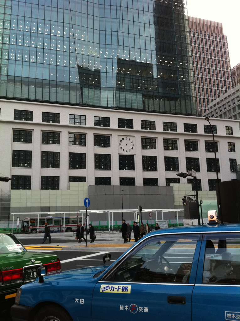 Eine Szene mit mehreren Autos, Holzplanken, einem Bus, Pfählen, Gebäuden und einigen Menschen, die stehen, mit lesbarem Text auf einem der Autos.