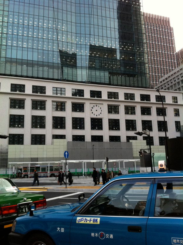 Eine Szene mit mehreren Autos, Holzplanken, einem Bus, Pfählen, Gebäuden und einigen Menschen, die stehen, mit lesbarem Text auf einem der Autos.