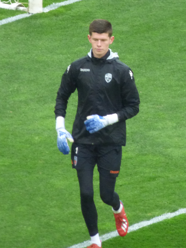 Ein Mann in einer schwarzen Jacke und blauen Handschuhen läuft auf einem Fußballfeld mit einem Tor im Hintergrund, trägt Fußballhandschuhe.