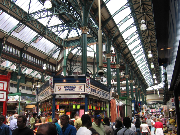 Ein belebter Markt mit Menschen, die durch Stände mit Waren, Schildern und hängenden Lichtern schlendern und eine lebendige Atmosphäre schaffen.