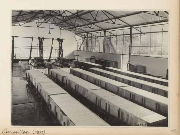 Ein Schwarz-Weiß-Foto einer Fabrik mit einem Zug auf den Schienen, umgeben von Maschinen, Masten, Lichtern und Glasfenstern, mit Text unten.