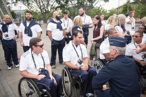 Eine Gruppe von Menschen im Rollstuhl, die vor einer Kulisse aus Bäumen, Pfählen, Gebäuden und einem klaren blauen Himmel miteinander sprechen, wobei einige Mützen und Brillen tragen und damit auf eine Teilnahme an den Paralympischen Spielen hindeuten.