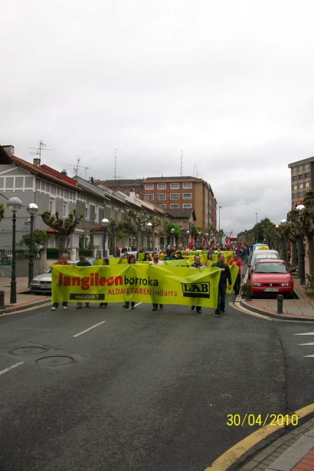Eine Gruppe von Menschen hält Schilder und marschiert auf einer Stadtstraße, mit Fahrzeugen, Polen und Bäumen auf den Gehwegen, Gebäuden im Hintergrund und einem bewölkten Himmel darüber.