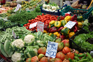 Ein belebter Bauernmarkt mit Körben voller Gemüse, darunter Tomaten, Paprika, Blumenkohl und grüne Bohnen, mit Preisschildern und einigen Menschen in der Nähe.