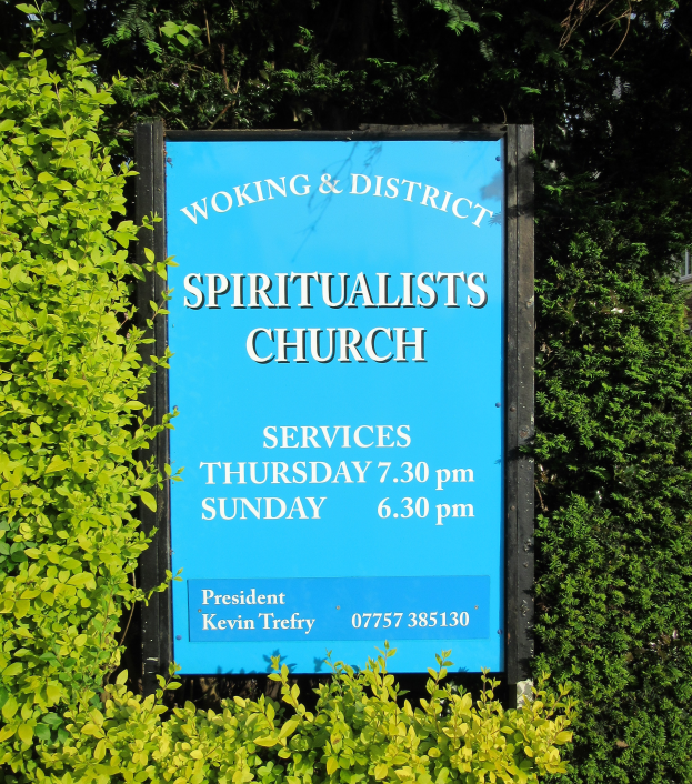 Ein Schild mit der Aufschrift "Woking & District Spiritualists Church" umgeben von Pflanzen und Bäumen, mit einem Gebäude im Hintergrund.