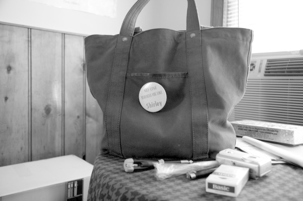 Eine Handtasche mit einer Kamera und einer Zigarettenbox darauf steht auf einem Tisch in einem Raum, mit einer Klimaanlage und einem Fenster im Hintergrund; die Tasche hat ein Etikett, auf dem steht "Keiner bedient Sie wie Shirley."