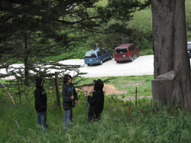 Drei Kinder in schwarzen Kleidern stehen auf einer grünen Fläche mit Fahrzeugen und Bäumen im Hintergrund.