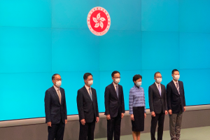 Eine Gruppe von Menschen mit Masken steht vor einer blauen Wand mit Logo, an einer Pressekonferenz in Hong Kong teilnehmend.