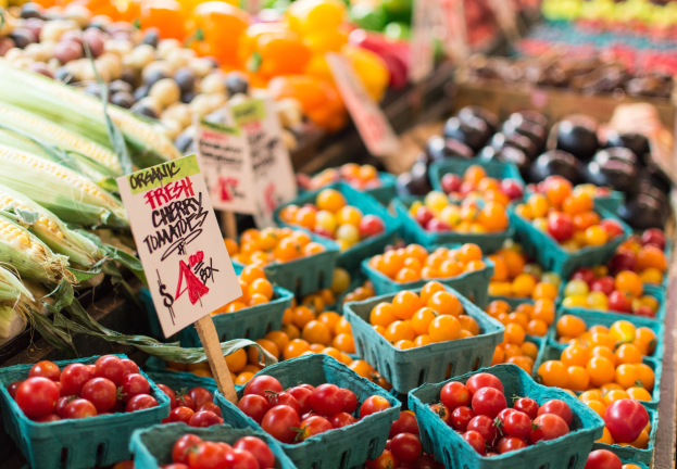 Frisches Obst und Gemüse wie Tomaten und Mais in Körben auf einem Bauernmarkt, mit Texttafeln im Hintergrund.