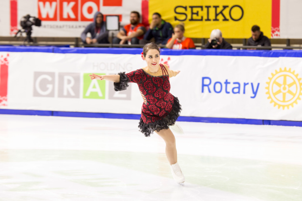 Eine Frau in einem roten und schwarzen Kleid läuft auf einer Eisfläche während der Weltmeisterschaften im Eiskunstlauf, umgeben von Zuschauern und einem Kameramann im Hintergrund.