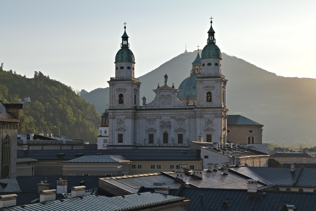 Panoramablick auf Salzburg, Österreich von einem Hügel aus, mit Gebäuden, Bäumen, einer prominenten Kathedrale, einem blauen und weißen Himmel und fernen Bergen.