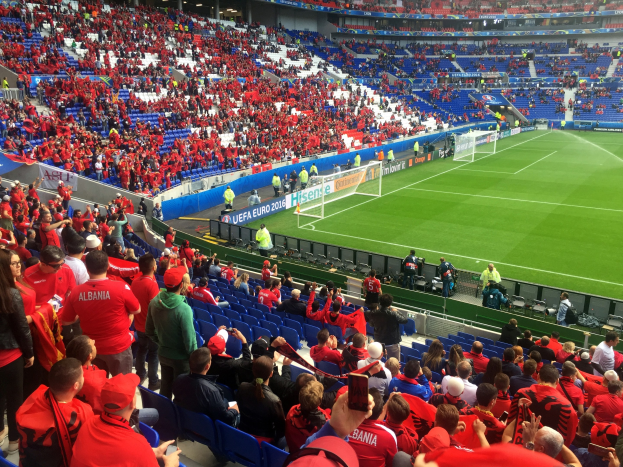 Eine große Menschenmenge in einem Stadion schaut einem Fußballspiel zu, mit Sitzplätzen, Stehplätzen, Bannern, einem Torpfosten und Deckenbeleuchtung.