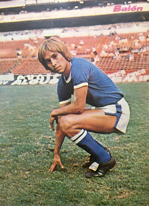 Ein Mann kniend auf einem Fußballfeld umgeben von Menschen auf Stühlen und Schildern mit Text.